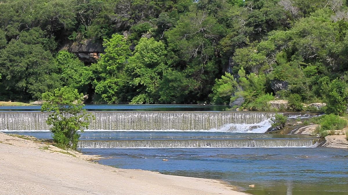 The San Gabriel River running clear and green over a low dam in Georgetown, Texas, with a limestone bluff and trees behind